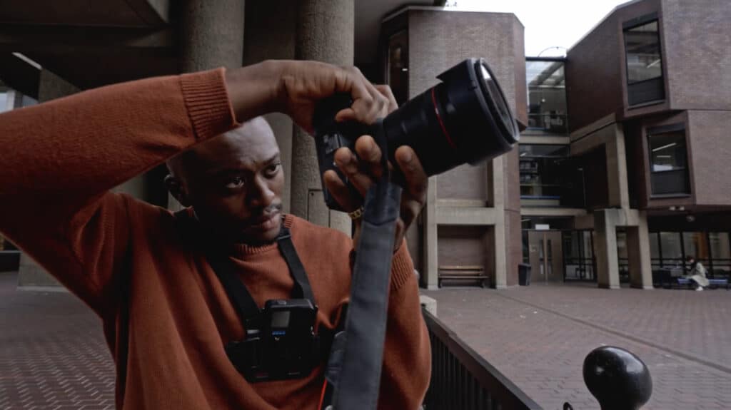 A man standing in a public square while holding a DSLR camera and looking at its screen.