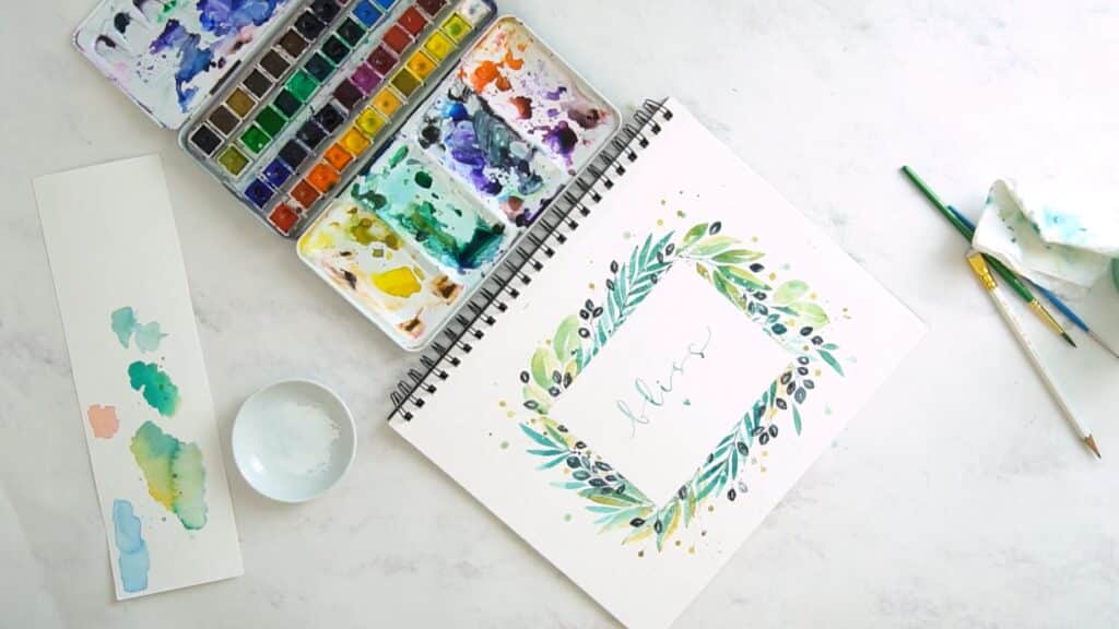 Spiral watercolor paper pad open to a watercolor painting of a botanical frame in blues, greens, and golds. Around the painting are a tray of watercolor paints, brushes, blotter paper and a bowl of water.