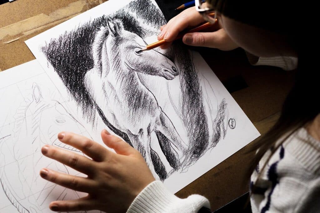An over-the-shoulder shot of a woman drawing on white paper with charcoal pencils. She is drawing an image of a baby horse, with a more rough sketch of the same drawing visible to the left.