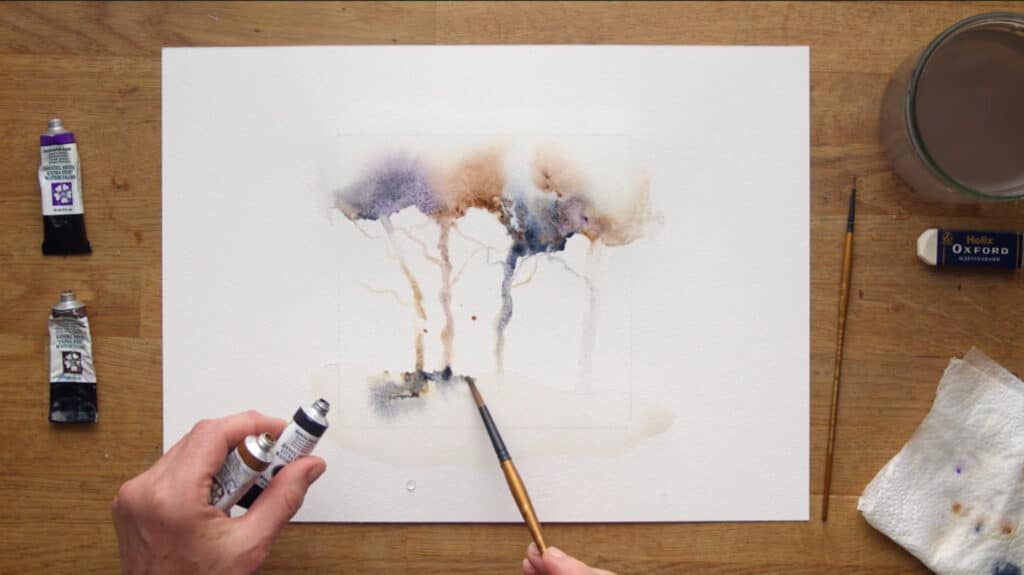 A woman’s hands using a paintbrush and tubes of watercolor paint to create a watercolor painting. The painting depicts abstract trees in shades of brown, purple and blue against a white background.