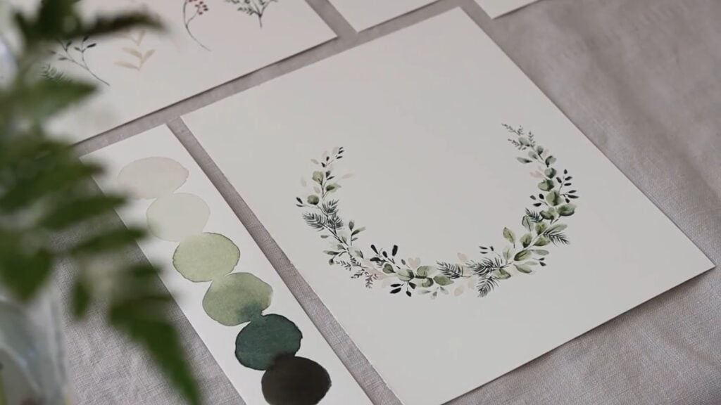 On a linen surface is a watercolor painting of a delicate, crescent-shaped winter wreath in a palette of creams, pale to dark greens and deep burgundy.