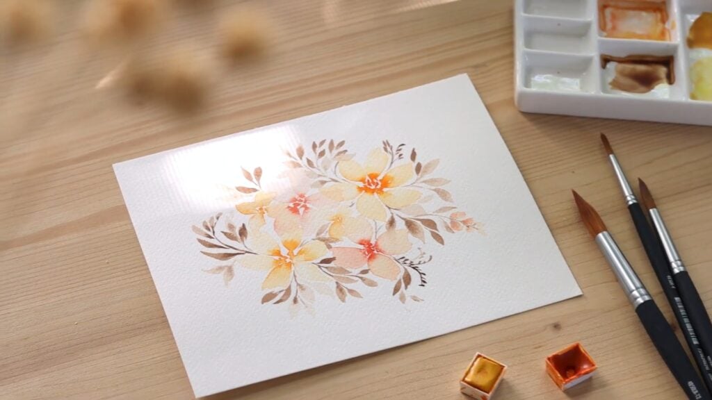 On a wooden surface is a watercolor painting of soft flowers and leaves in pale oranges, pinks and browns. Next to the painting are watercolor paints, brushes and a paint tray.