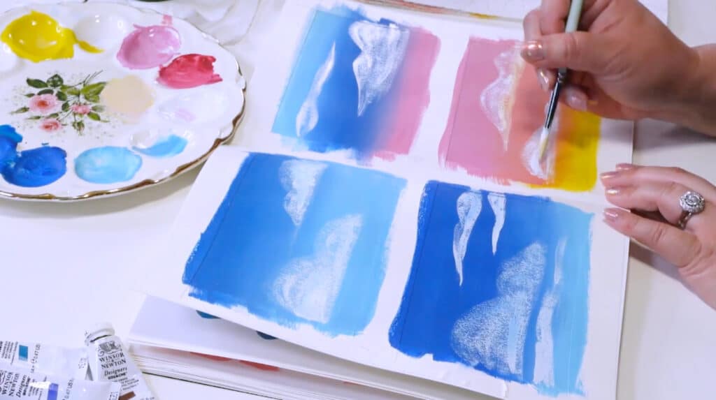 A woman’s hands painting white clouds over gradient blue, pink and yellow backgrounds.