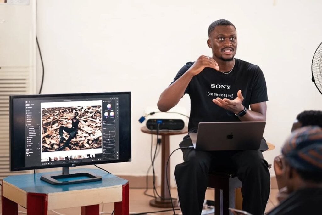 A man sitting in front of a class of people. He has a Macbook laptop on his lap. A monitor facing the students displays photo editing software he is using to demonstrate editing techniques. 