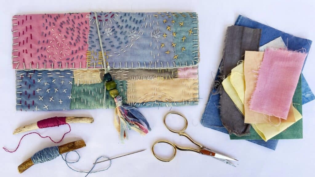 A multicolored tri-fold pouch decorated with visible stitches in various colors of thread. The pouch lies on a white tabletop next to scissors, thread, and fabric scraps.