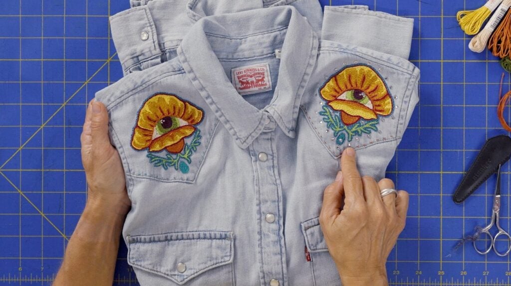 A person’s hands holding a light blue denim vest over a dark blue cutting mat. The front panels of the vest have been decorated with two embroidered orange flowers.