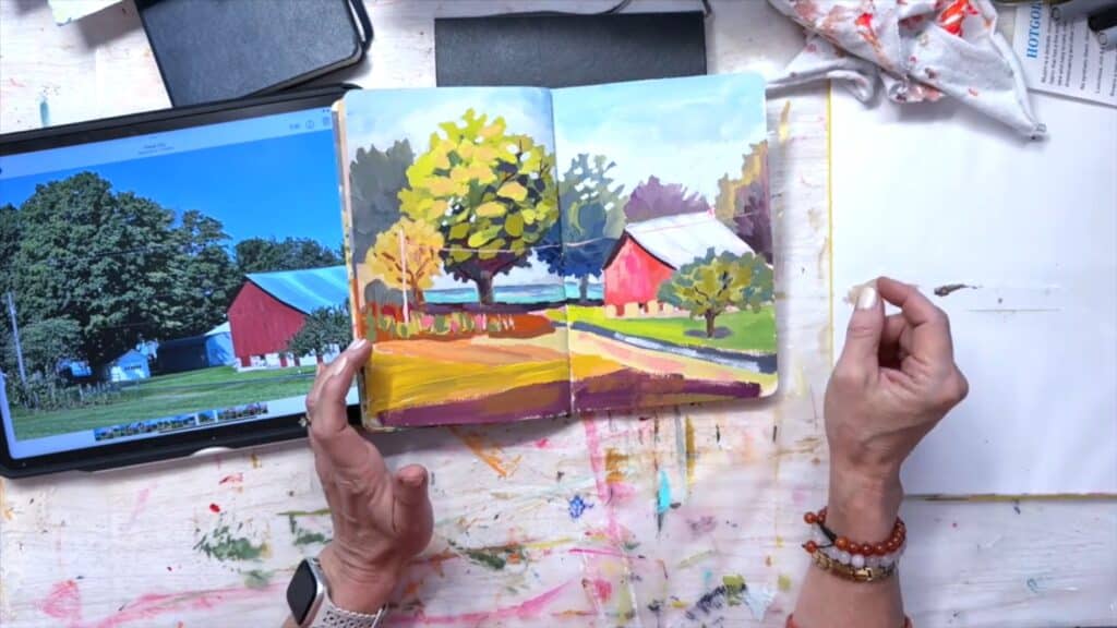Sketchbook open to a rural barn landscape painting, surrounded by a digital photo of the same scene, various art materials and the artist's hands