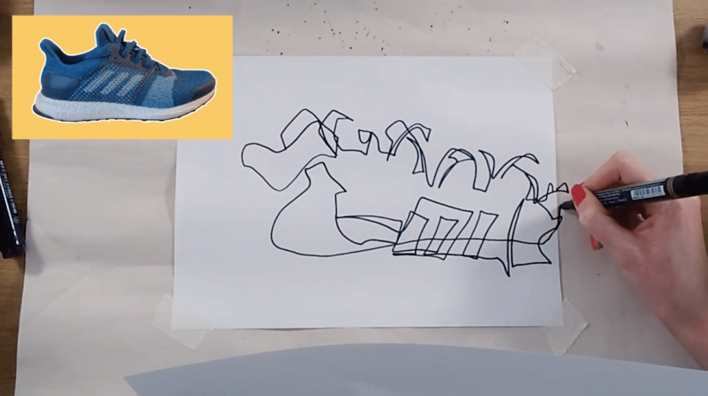 A white sheet of paper is taped to a bigger white piece of paper on a table. A hand holds a black marker, drawing an abstract shoe. The inspiration photo for the shoe drawing is located in the top right corner and features a blue sneaker on a yellow background. 