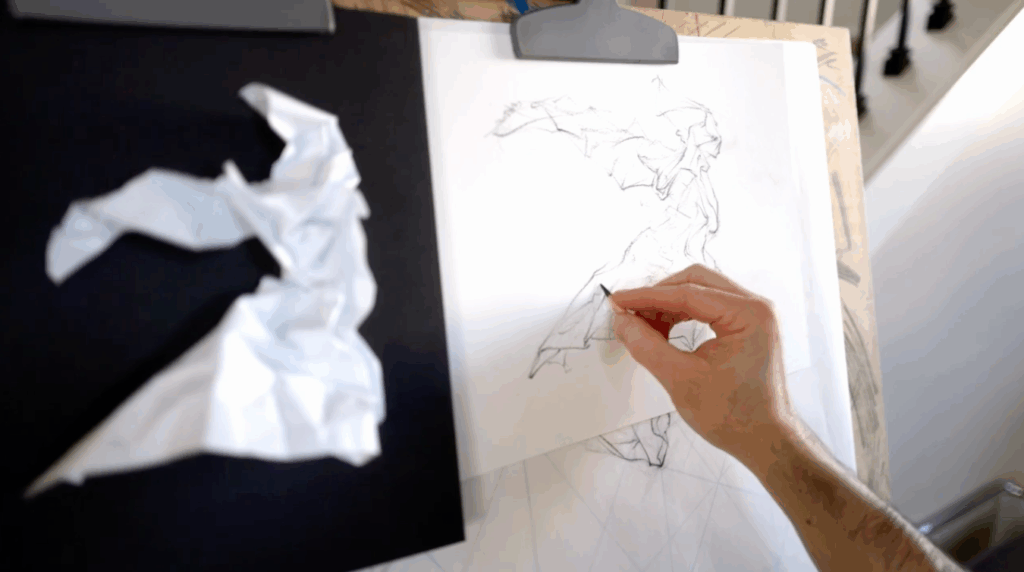 A few pieces of paper are clipped to a big board. The first piece of paper is black and has a crumpled-up piece of white printer paper pinned to it. The second piece of paper is white and features a sketch that mimics the form of the crumpled-up printer paper. A hand holding a pencil is adding shading to the sketch. 