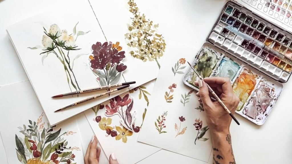 Desk covered in pages of wildflower watercolors in muted oranges, burgundies, greens and goldens, with the artist’s hand and palette nearby