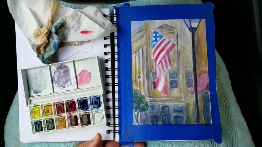 An open sketchbook and a palette of watercolors sitting on a person’s lap. Its page is filled with a watercolor painting of an American flag hanging in front of an ornate stone building.