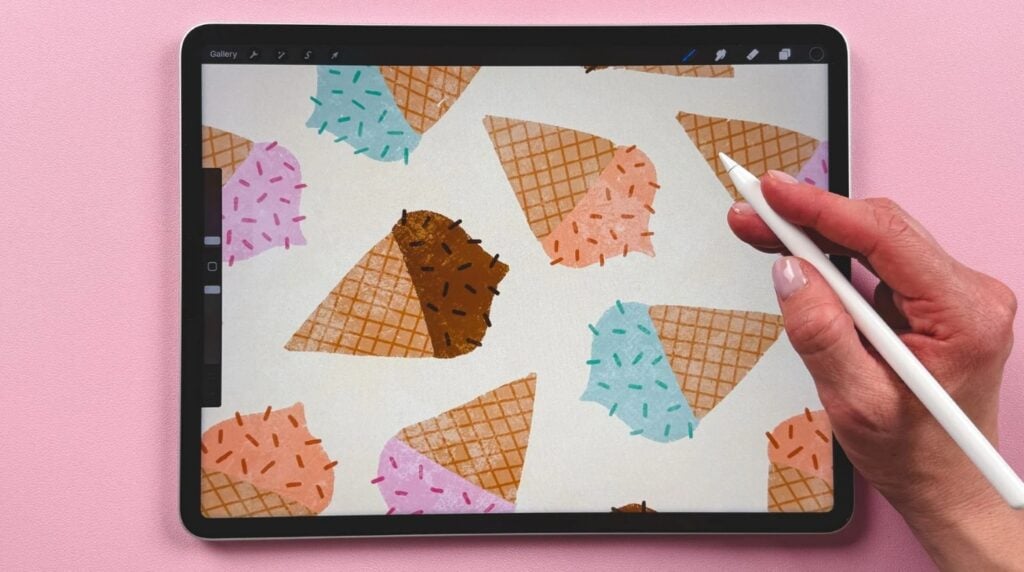 A person using an Apple Pencil to draw on an iPad that’s resting on a pink background. The iPad’s screen is filled with a pattern consisting of colorful ice cream cones.
