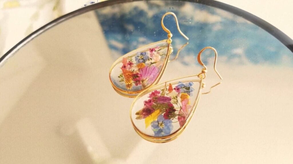 A pair of gold teardrop earrings lying on a mirrored surface. The drop-shaped centers of the earrings are filled with pressed flowers suspended in clear resin.
