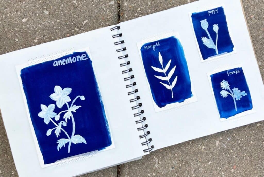 A sketchbook lying open on a concrete surface. Its pages are filled with white silhouettes of flowers against deep blue backgrounds.