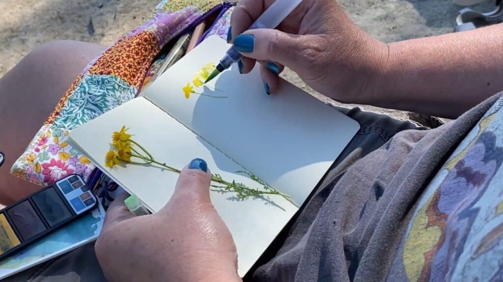 A person using watercolors to paint a yellow flower in a notebook while outdoors.
