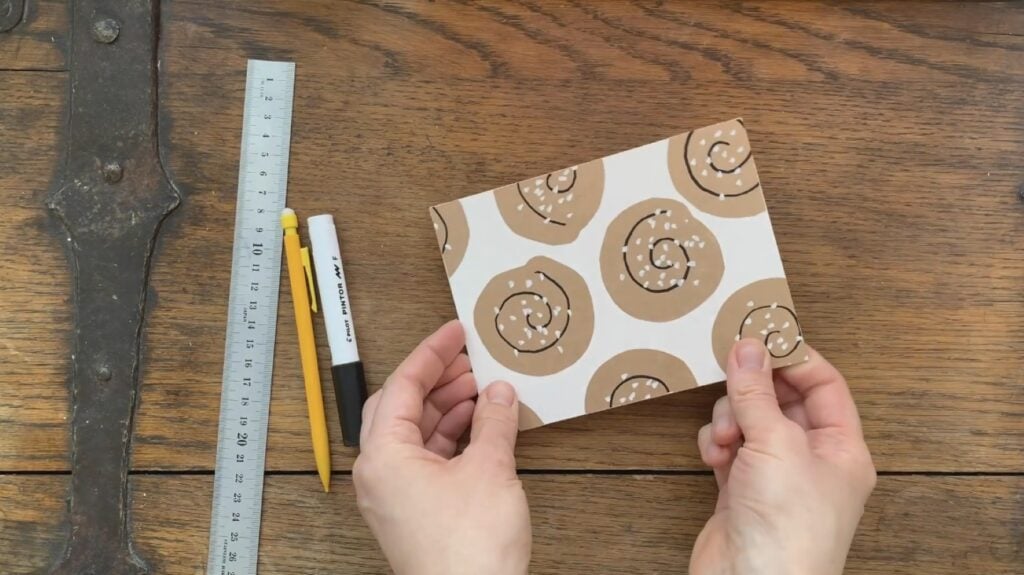 A person’s hands holding a postcard over a brown wooden surface. The postcard is white and features brown cinnamon roll-like designs decorated with black spirals and white dots.