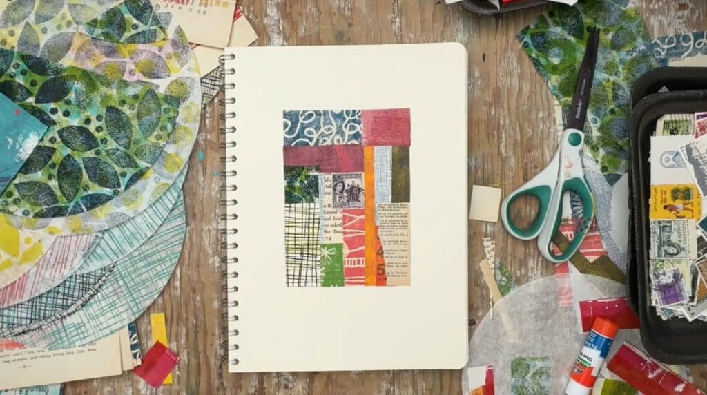 A sketchbook laying on a wooden table. Its page is decorated with a patchwork collage made of scraps of patterned paper.
