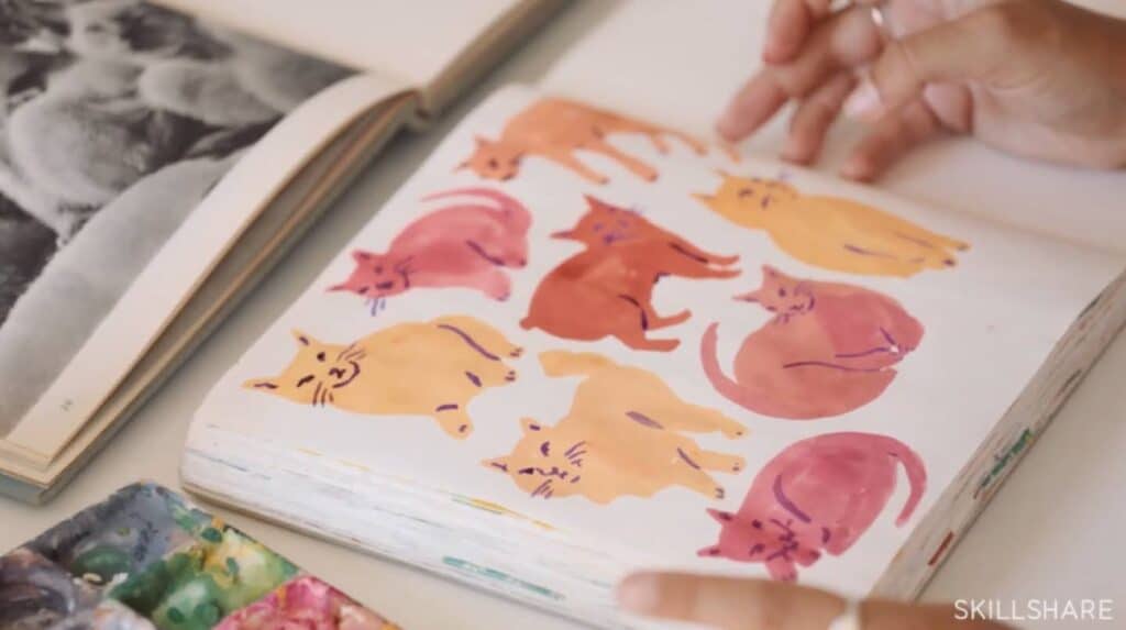 A person’s hands resting near a sketchbook laying open on a table. The page is filled with pink and orange watercolor illustrations of cats in various poses.