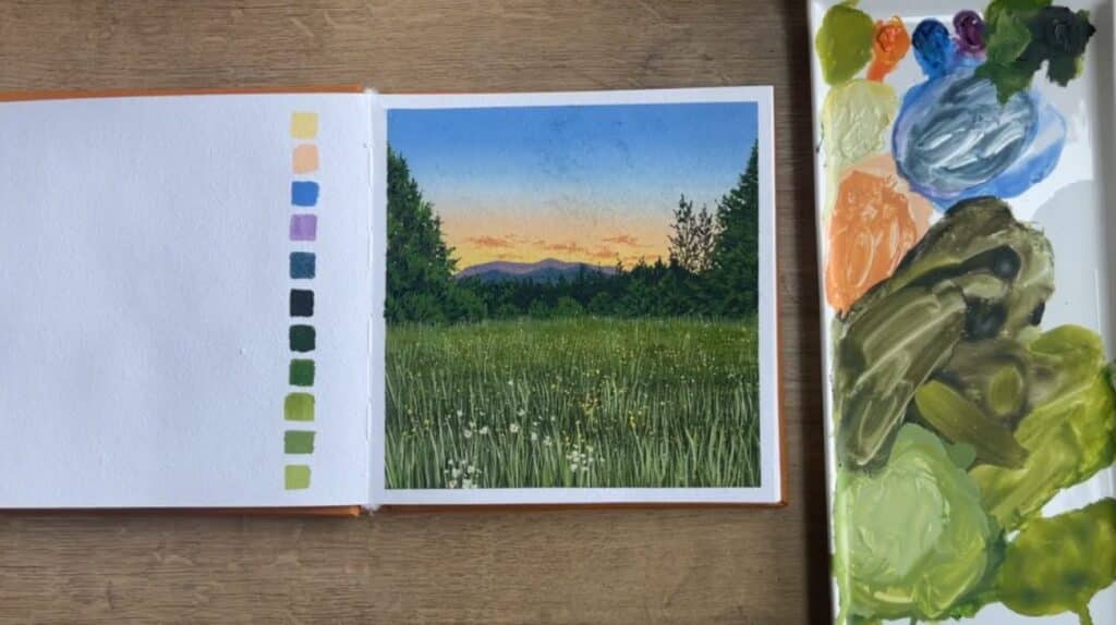 A sketchbook laying open on a table next to a gouache paint palette. Its page is filled with a painting of a lush green meadow in the foreground and silhouetted mountains in the background.