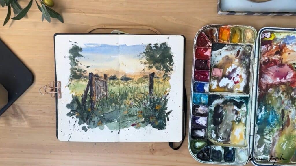 A sketchbook laying open on a table alongside a watercolor paint palette. The sketchbook’s pages are filled with a painting of a wooden gate in the foreground and a sunset in the background.