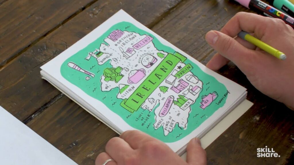 A person’s hands drawing a brightly colored map of Ireland on a white piece of paper. The map is decorated with several small doodles alongside labels of major areas.