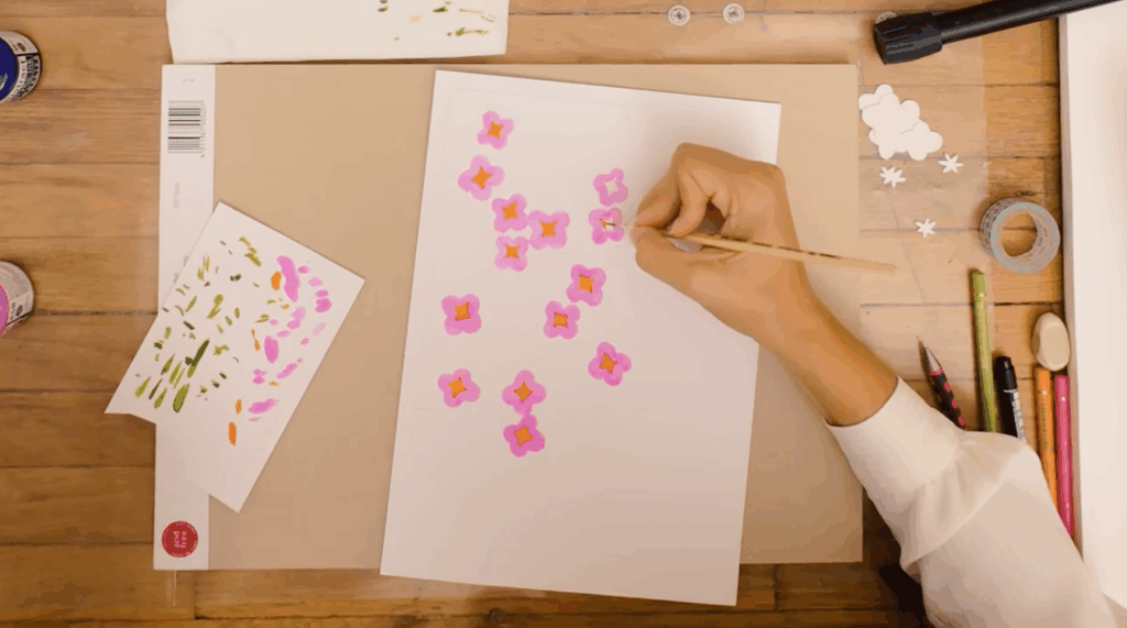 A piece of white paper covered in hand-painted pink flowers sits on a wooden desk surrounded by art materials like washi tape, paint and pencils. A hand holding a paintbrush adds a touch of orange paint to the middle of one of the pink flowers. 