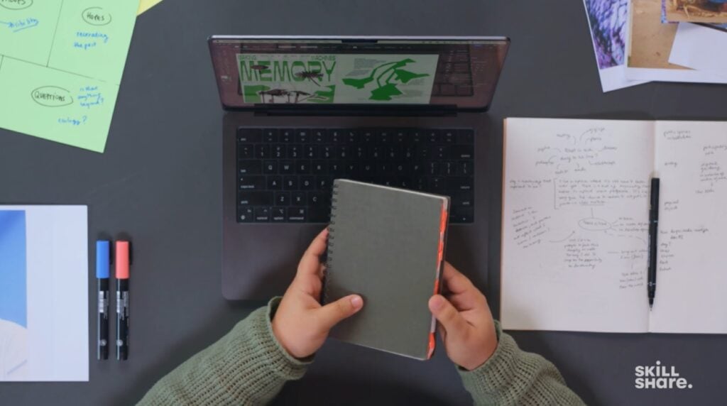 Alt: An overhead view of a person’s hands holding a small notebook over an open Macbook computer.