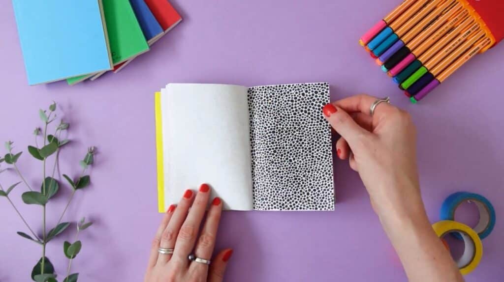 A person’s hands opening a sketchbook on a purple tabletop. The sketchbook is filled with a full-page doodle of black dots in various sizes.