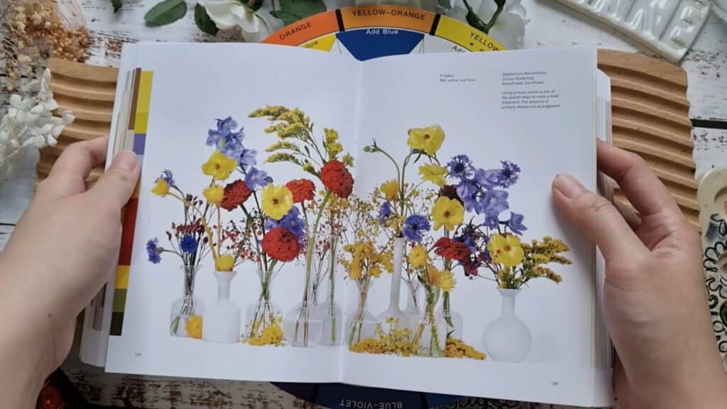 Book open to a page with several vases of flowers in reds, yellows, and blues, laying on a color wheel and wooden work surface, surrounded by various tools and botanicals