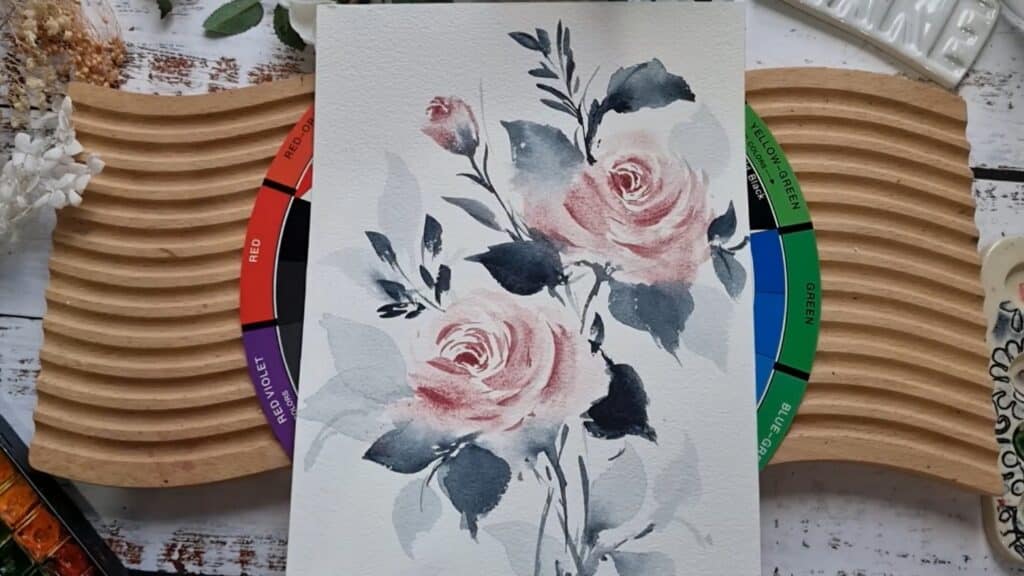 Painting of pale pink roses with gray-blue stems and leaves, laying on a color wheel and wooden work surface, surrounded by various tools and botanicals