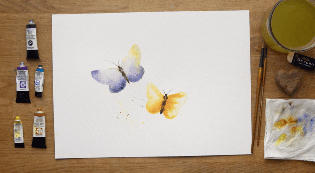 A watercolor painting of two butterflies sits on a wooden table next to five tubes of paint, two paint brushes, a paper towel, an eraser and a cup of water. 