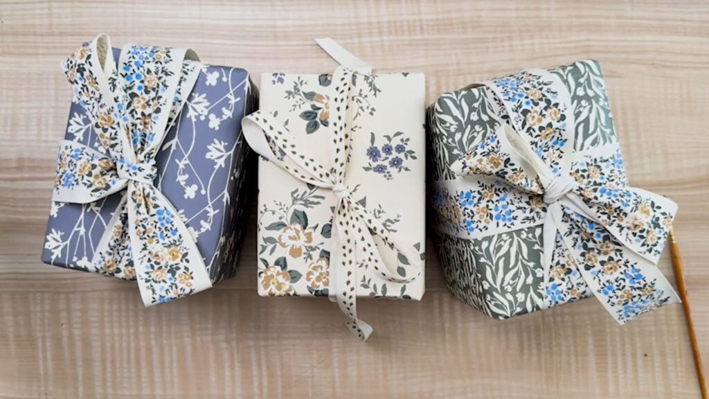 Three wrapped gifts, all in designer paper and ribbons featuring delicate florals in blues, golds, greens and cream, sitting on a wooden surface.