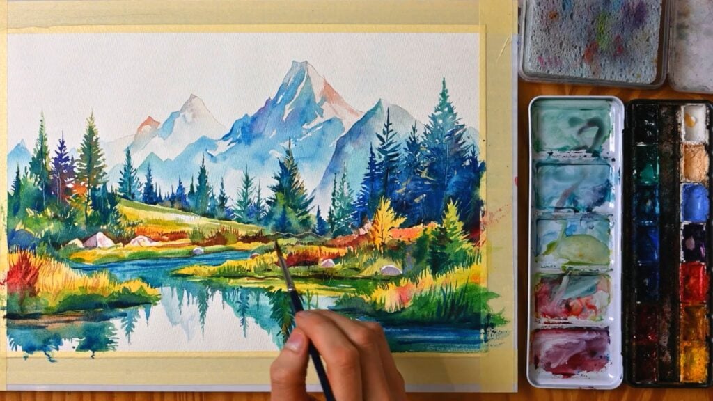 Artist adding the finishing touches to a colorful mountain scene in watercolor, with open paints and mixing trays opened just to the right