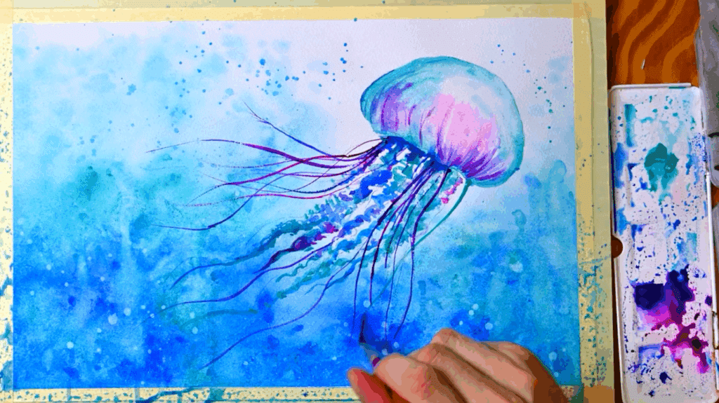 A watercolor painting of purple and blue jellyfish sits on a wooden table next to a palette of watercolor paint. A hand holding a paintbrush adds a stroke of blue to the canvas.