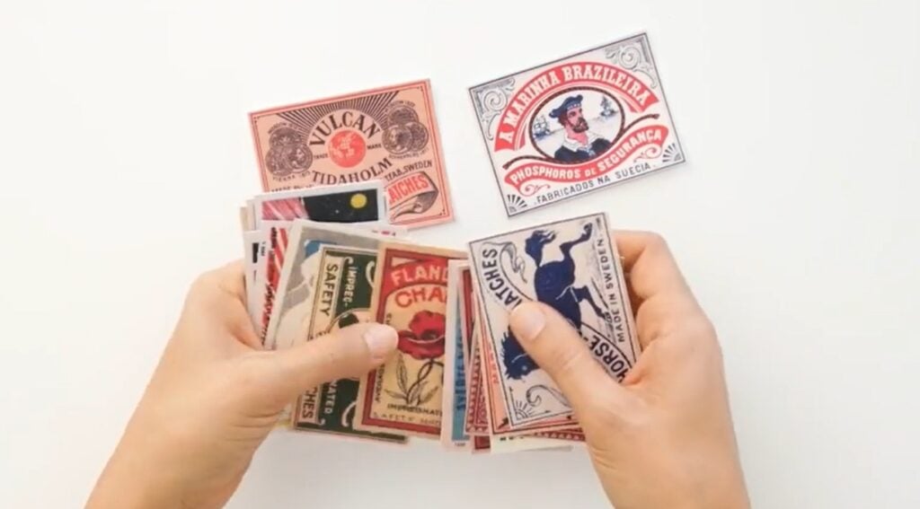 Pair of hands holding and sorting a stack of cards featuring vintage matchbox label art, most of which are in black, white, reds and sepia tones