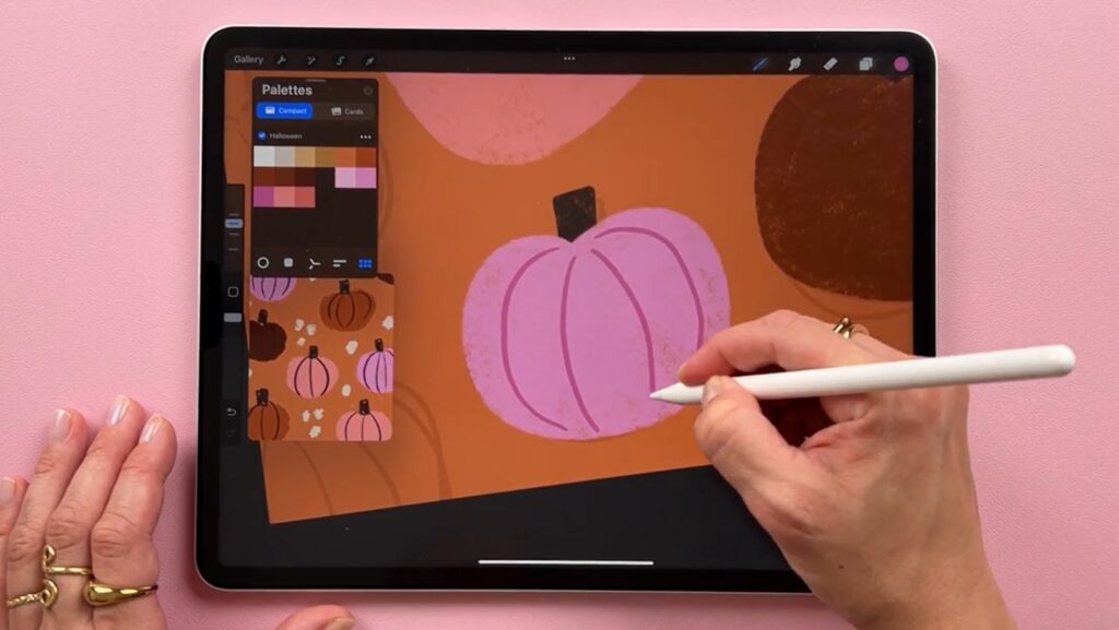 iPad with an open Procreate project, featuring little pink, orange, and brown pumpkins against an orange background, and the artist’s hand adding some finishing touches while referencing the color palette.