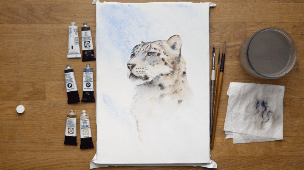 A watercolor painting of a snow leopard sits on a wooden table next to six tubes of paint, three paint brushes, a paper towel and a cup of water. 