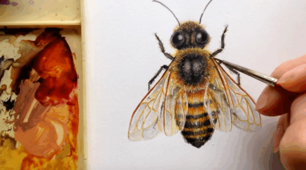 A watercolor painting of a bumblebee sits on a flat surface next to a paint palette. A hand holding a paintbrush adds strokes of yellow to the painting. 