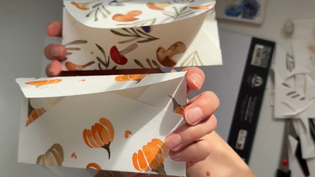  A person’s hands holding two envelopes. One is decorated with a watercolor pattern of orange pumpkins, and one is decorated with a watercolor pattern of mushrooms and leaves. 