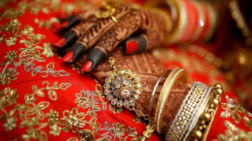 An Indian bride’s hands resting on her bright red dress. The dress is decorated with gold embroidery, and her hands are decorated with sparkling bracelets and rings. 