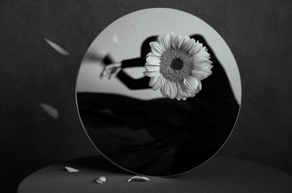 A black and white photo of a round mirror. In the mirror’s reflection a dancing woman in a black dress is visible. A sunflower covers the woman’s face.