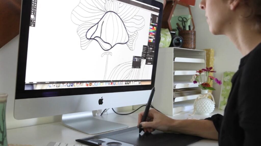 A woman sitting at a desk and using a drawing tablet to illustrate a flower on her computer screen.