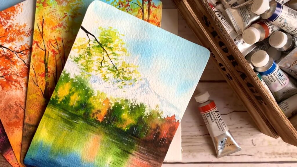 Fanned out stack of watercolor autumn landscapes next to a crate of paint tubes atop a wooden work surface