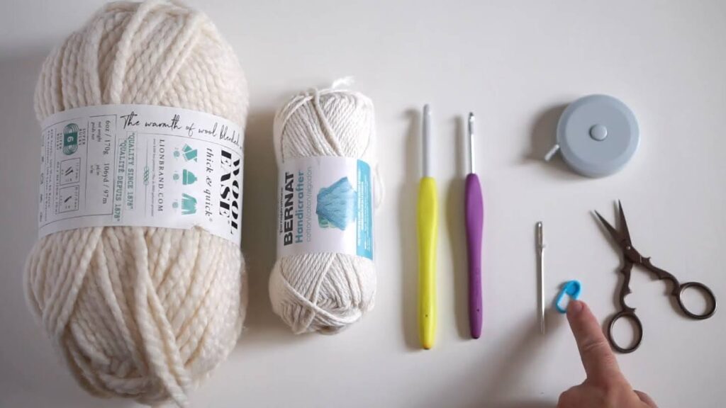From left to right on a white work surface: a skein of bulky, beige yarn, a skein of finer, off white yarn, two crochet hooks in different lengths, a yarn needle, stitch marker, measuring tape and scissors