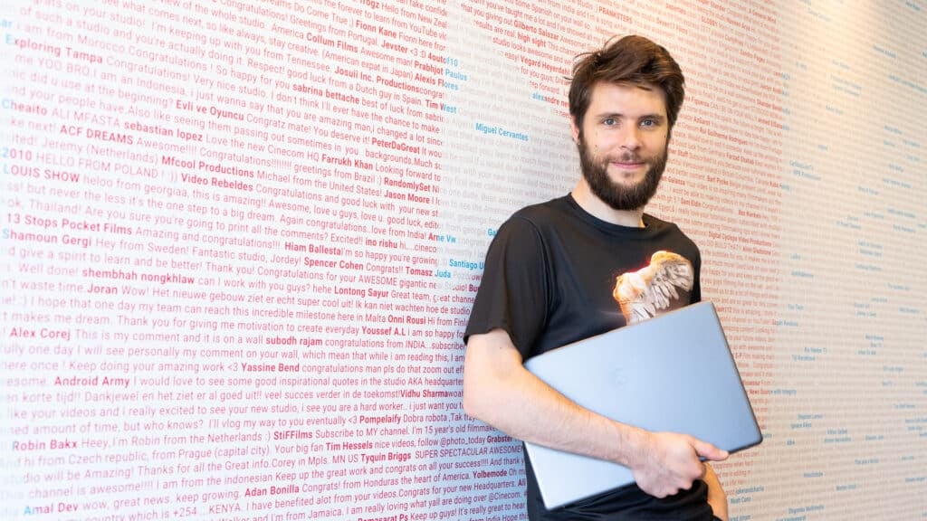 A man holding a laptop leans against a wall with text covering it.