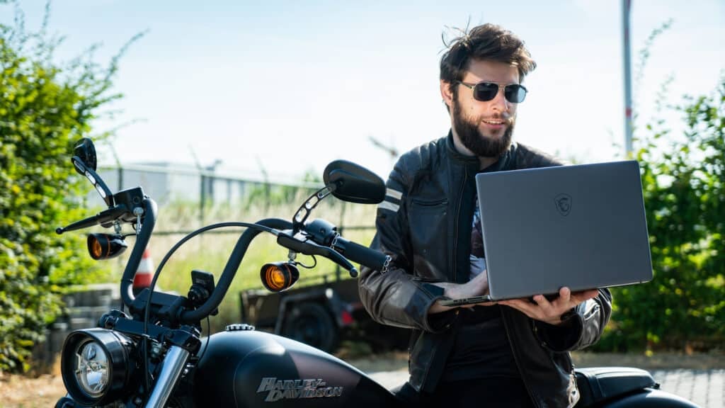 A man wearing shades and a leather jacket sits on a Harley Davidson motorcycle. He is holding a laptop.