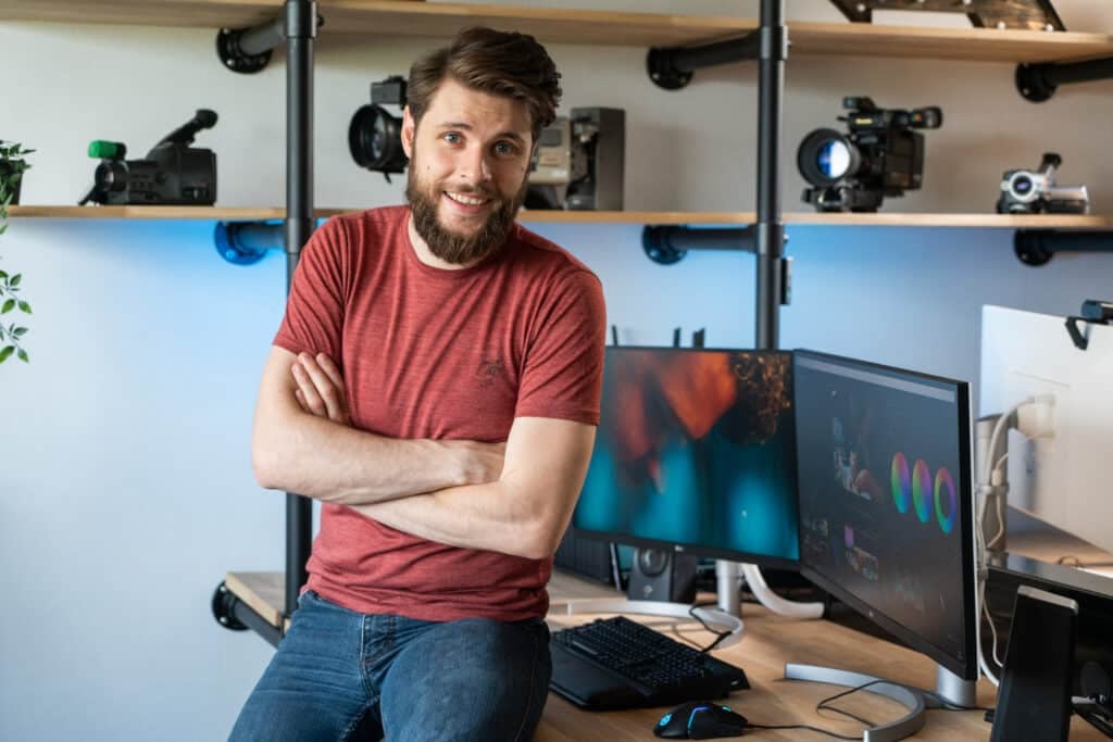 Photo of a man in jeans and a red t-shirt perched on his desk and smiling at the camera. Behind him are monitors featuring video editing software and camera equipment on shelves.