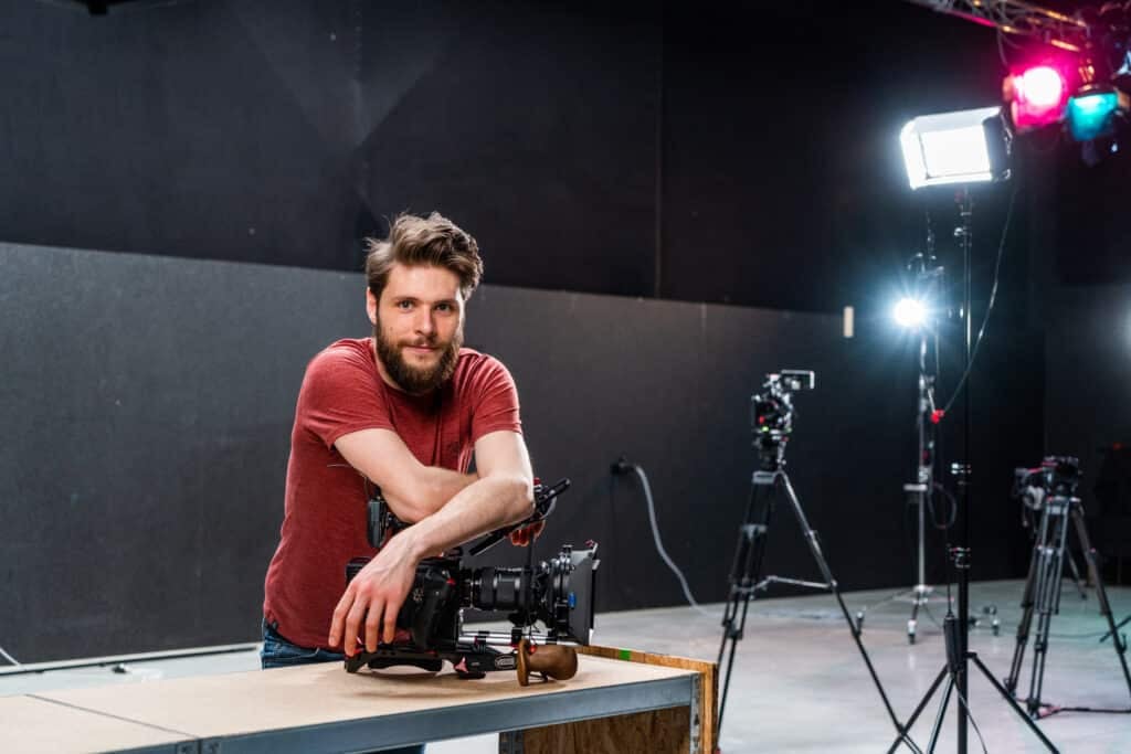 A man leans on a camera in a film studio. Behind him is scattered professional studio lighting equipment.
