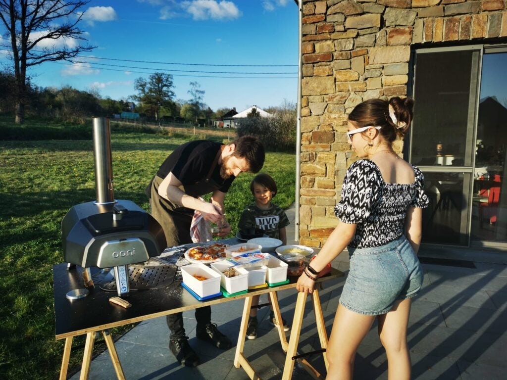 An outside shot of 3 people gathered around a table scattered with pizza toppings and a pizza oven