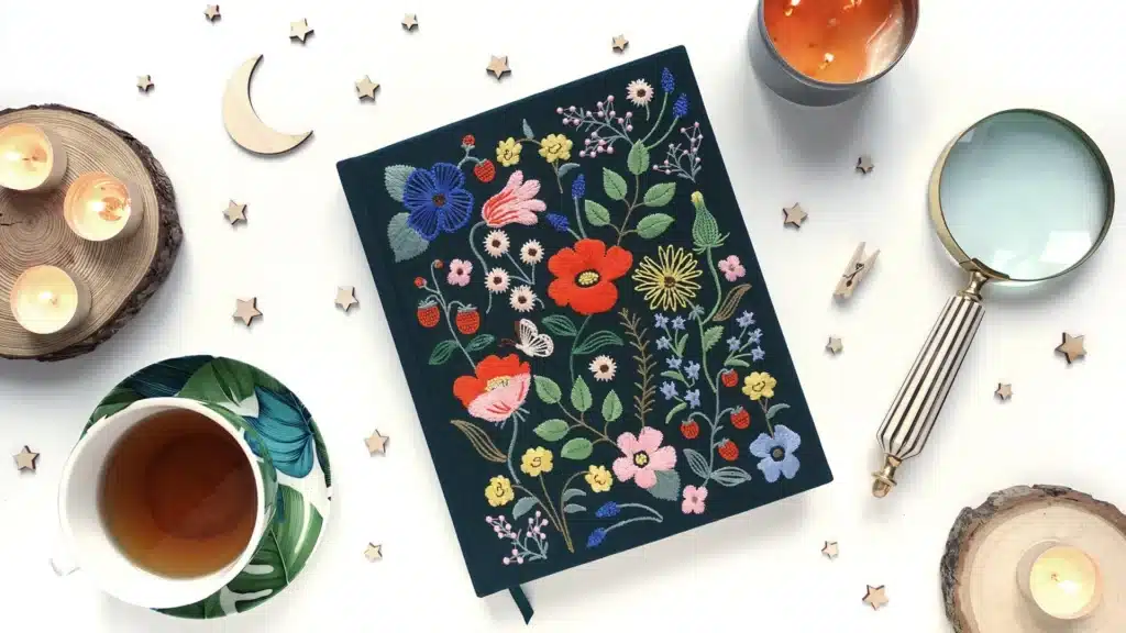 Overhead shot of a journal featuring a floral cover and surrounded by cozy objects such as a mug of tea, candles, and a magnifying glass